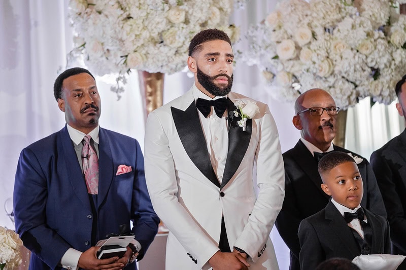 A wedding scene with a groom in a white tuxedo and black bow tie, adhering to the three-button suit rule, standing with groomsmen in black suits. The groom, solemn and