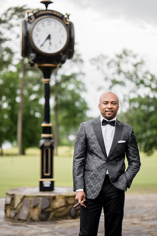 A man in a stylish one-button gray tuxedo with a black bow tie stands confidently in front of an ornate outdoor clock post in a park, with a blurred background featuring green trees and grass