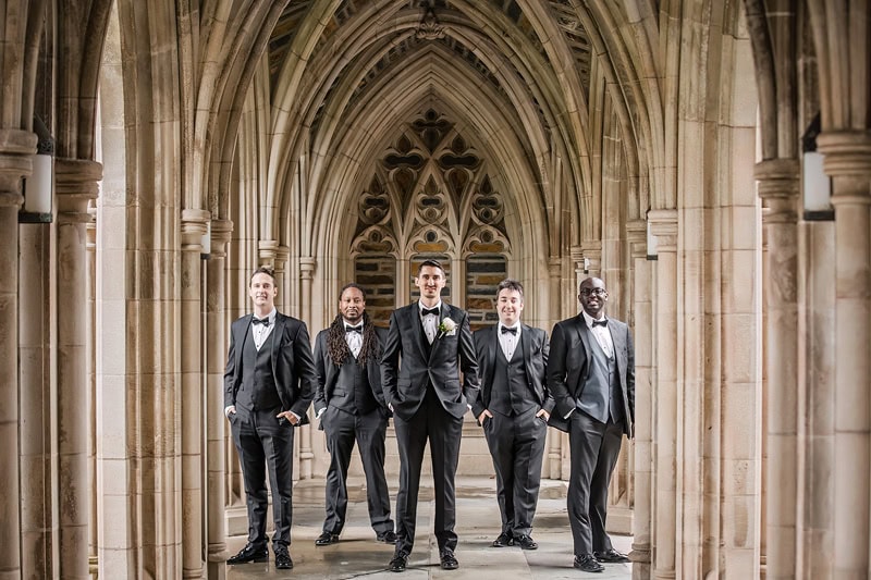 Five men in tuxedos stand in a row under the ornate arches of a gothic-style stone corridor, demonstrating how to button their jackets, and smiling slightly towards the camera. The