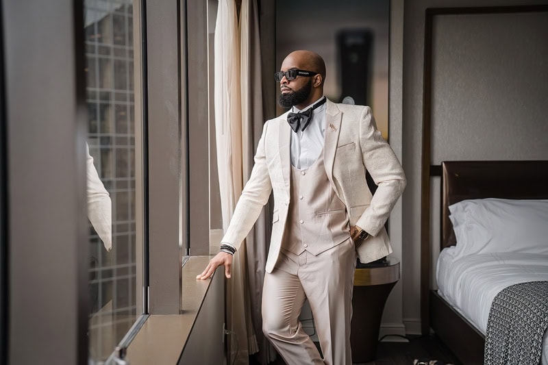 A stylish man in a two-button beige suit and bow tie stands by a window in a modern hotel room, looking away thoughtfully. He wears dark sunglasses and a well-groomed beard, positioned