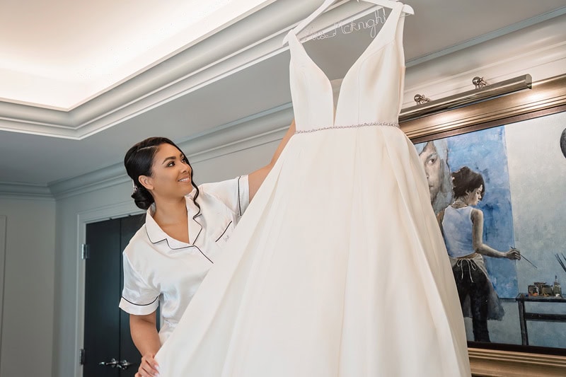 A woman in a white blouse smiles while adjusting a hanging white wedding dress in the Grand Bohemian Hotel Charlotte, adorned with elegant decor and a painting of a ballerina on the wall.