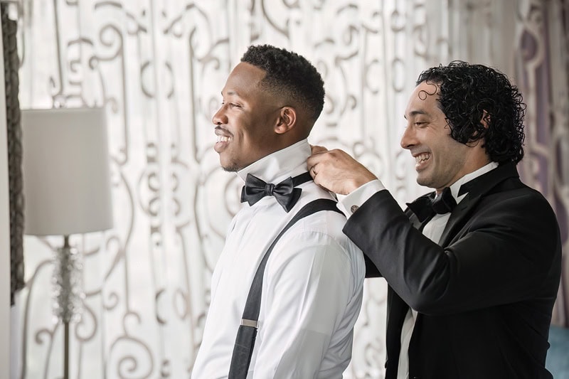 Two men in tuxedos smile while standing indoors at the Grand Bohemian Hotel Charlotte, one of Asian descent adjusts the other's bow tie, who is of African descent. They appear joyful