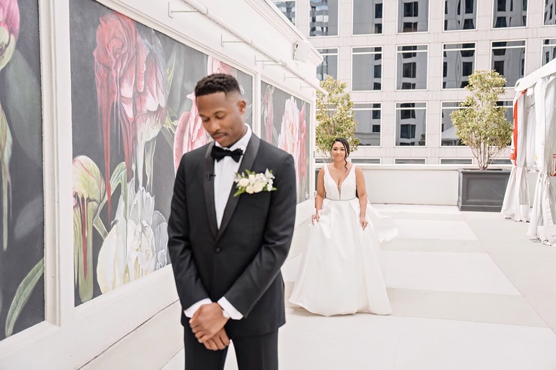 A groom in a black tuxedo looks down thoughtfully on a rooftop decorated with floral designs at the Grand Bohemian Hotel Charlotte, as a bride in a white gown approaches from behind, with