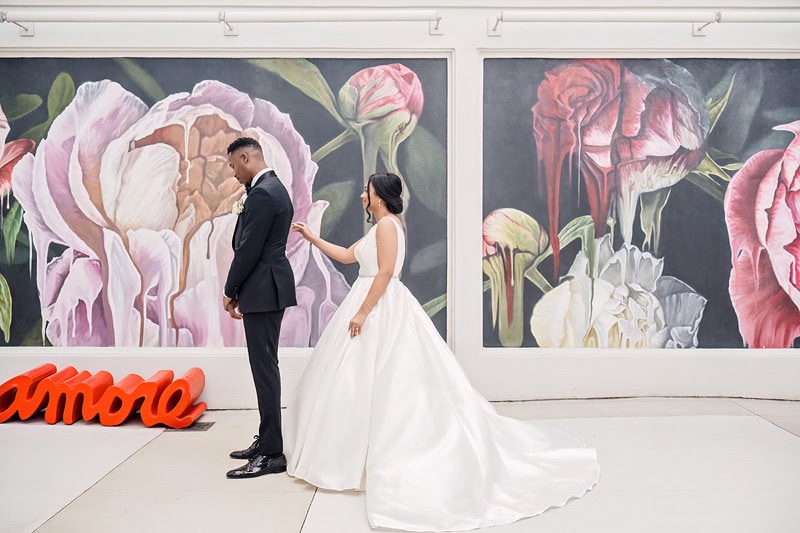 A bride and groom, dressed in elegant wedding attire, hold hands facing away from the camera against a backdrop of a large, vibrant floral painting at the Grand Bohemian Hotel Charlotte, with a neon