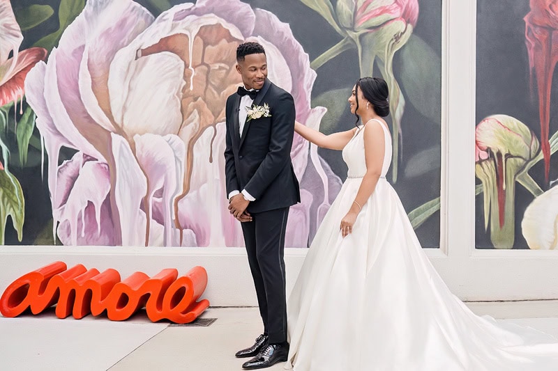 A joyful couple in wedding attire share a moment at the Grand Bohemian Hotel Charlotte, standing in front of a large floral mural with the word "amore" in red letters at the bottom.