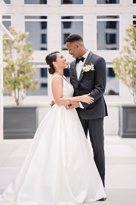 A bride in a white gown and a groom in a black tuxedo lovingly gaze at each other, standing in front of the Grand Bohemian Hotel Charlotte, adorned with geometric patterns and small