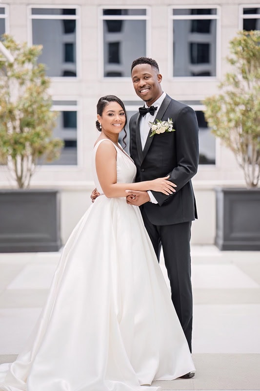 A smiling bride and groom stand closely, posing outdoors at the Grand Bohemian Hotel Charlotte. The bride wears a white sleeveless wedding gown with a long train, and the groom is in a classic