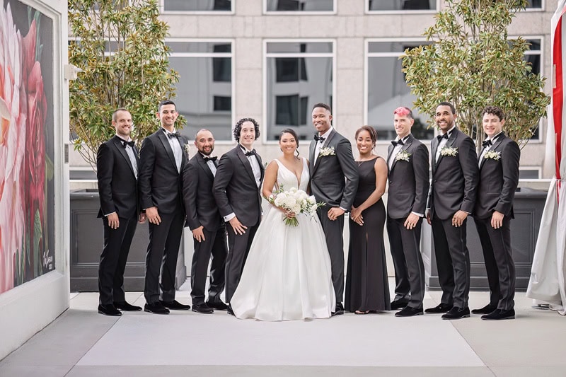 A wedding party outside the Grand Bohemian Hotel Charlotte, featuring a bride in a white gown holding a bouquet, flanked by four bridesmaids in black dresses and five groomsmen in black suits