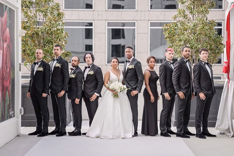 A wedding group stands on the rooftop of Grand Bohemian Hotel Charlotte, featuring eight people - seven men in black tuxedos and a woman in a white gown, holding a bouquet. They