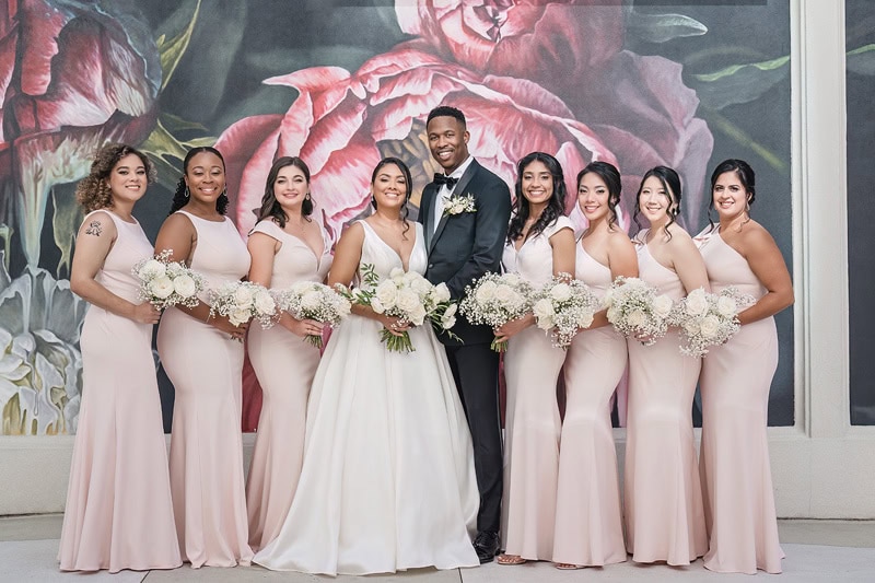 A smiling groom in a dark suit stands surrounded by seven bridesmaids in flowing pink dresses, each holding a bouquet, in front of a large floral mural at the Grand Bohemian Hotel Charlotte.