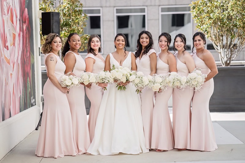 A bride in a white dress stands with seven bridesmaids in matching pale pink dresses, all holding bouquets. They smile joyfully in front of the Grand Bohemian Hotel Charlotte, adorned with a