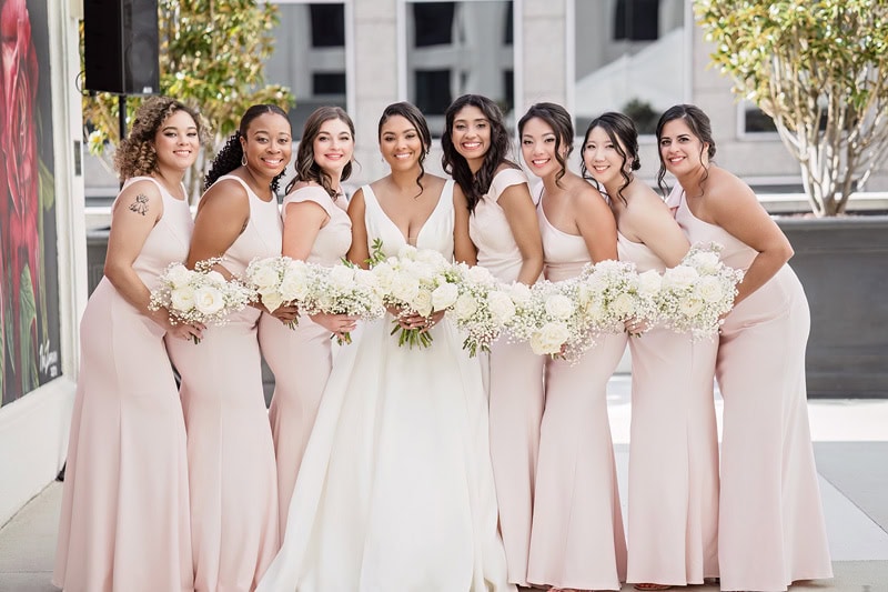 A diverse group of eight bridesmaids and a bride posing joyfully outside the Grand Bohemian Hotel Charlotte. The bridesmaids wear matching pale pink dresses and hold white floral bouquets; the bride in