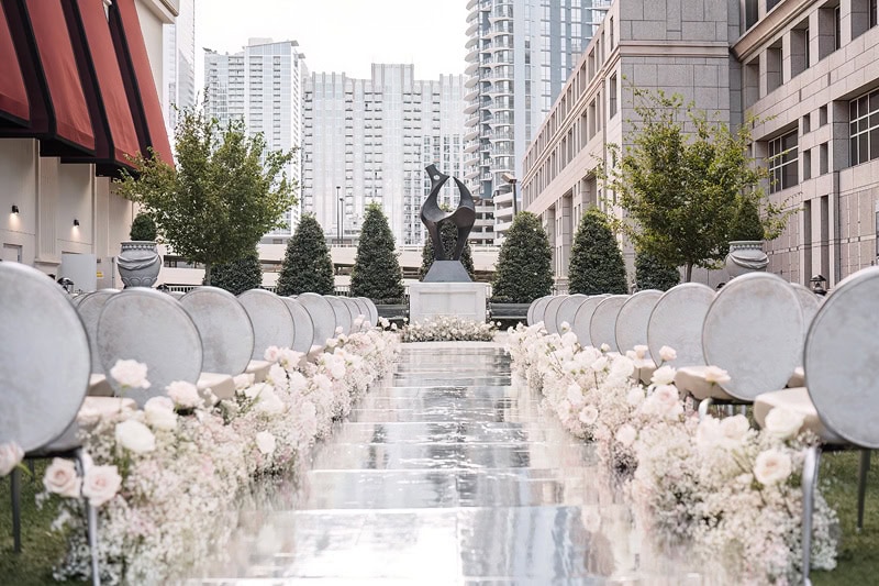 An outdoor wedding aisle at the Grand Bohemian Hotel Charlotte lined with white chairs and light pink roses, leading to a sculpture at the end, set against a backdrop of high-rise buildings.