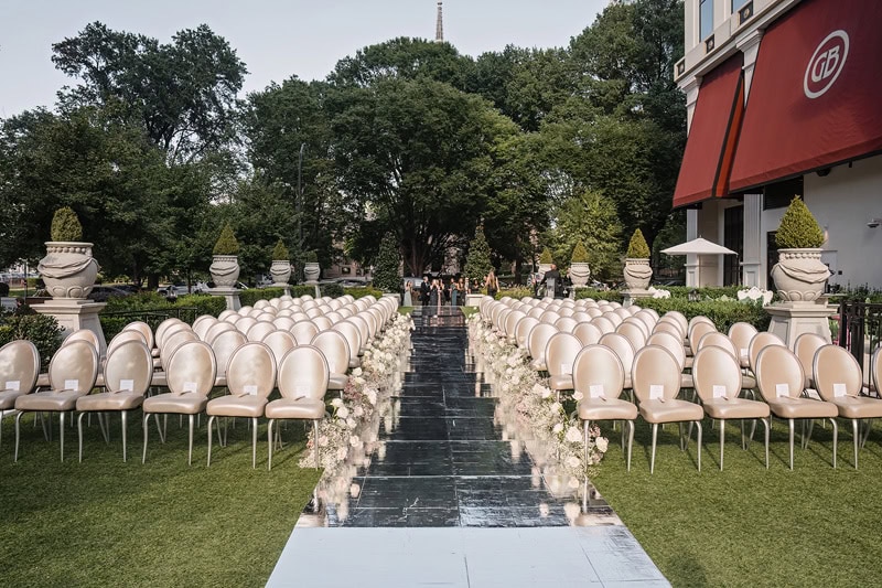 An outdoor wedding setup at Grand Bohemian Hotel Charlotte featuring rows of white chairs on either side of a black aisle runner, bordered by floral arrangements. Trees and a partial view of a building with a