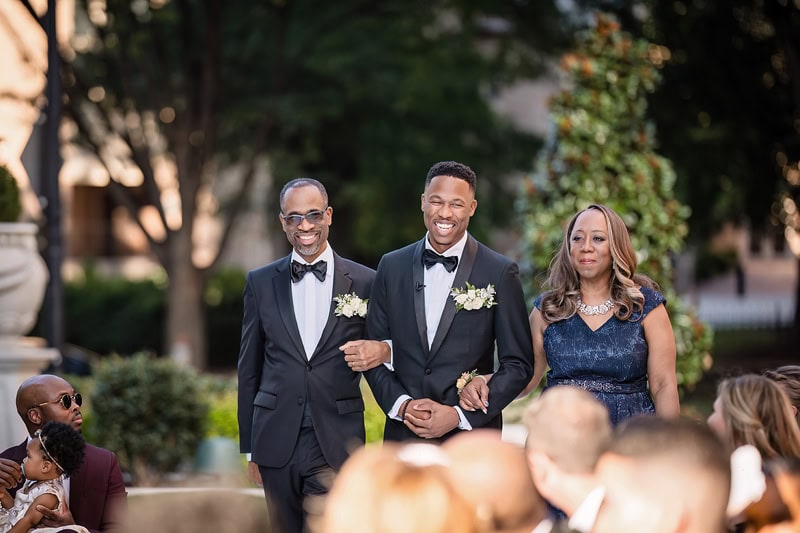 A joyful wedding scene outdoors at the Grand Bohemian Hotel Charlotte with a groom walking down the aisle, flanked by an older man and a woman, all smiling broadly in formal attire. Guests are