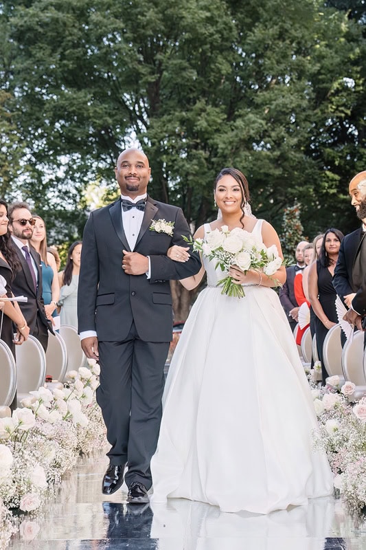 A bride and groom walk down the aisle hand-in-hand, smiling, at their outdoor wedding ceremony at the Grand Bohemian Hotel Charlotte. The groom is in a black tuxedo and the bride