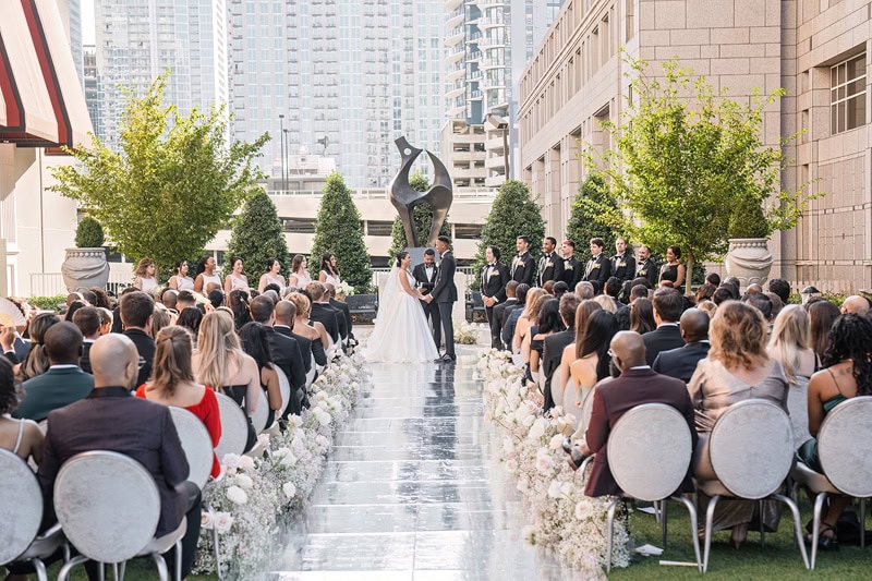A wedding ceremony outdoors at the Grand Bohemian Hotel Charlotte with a bride and groom standing at the altar, surrounded by guests seated in rows and urban skyscrapers in the background.