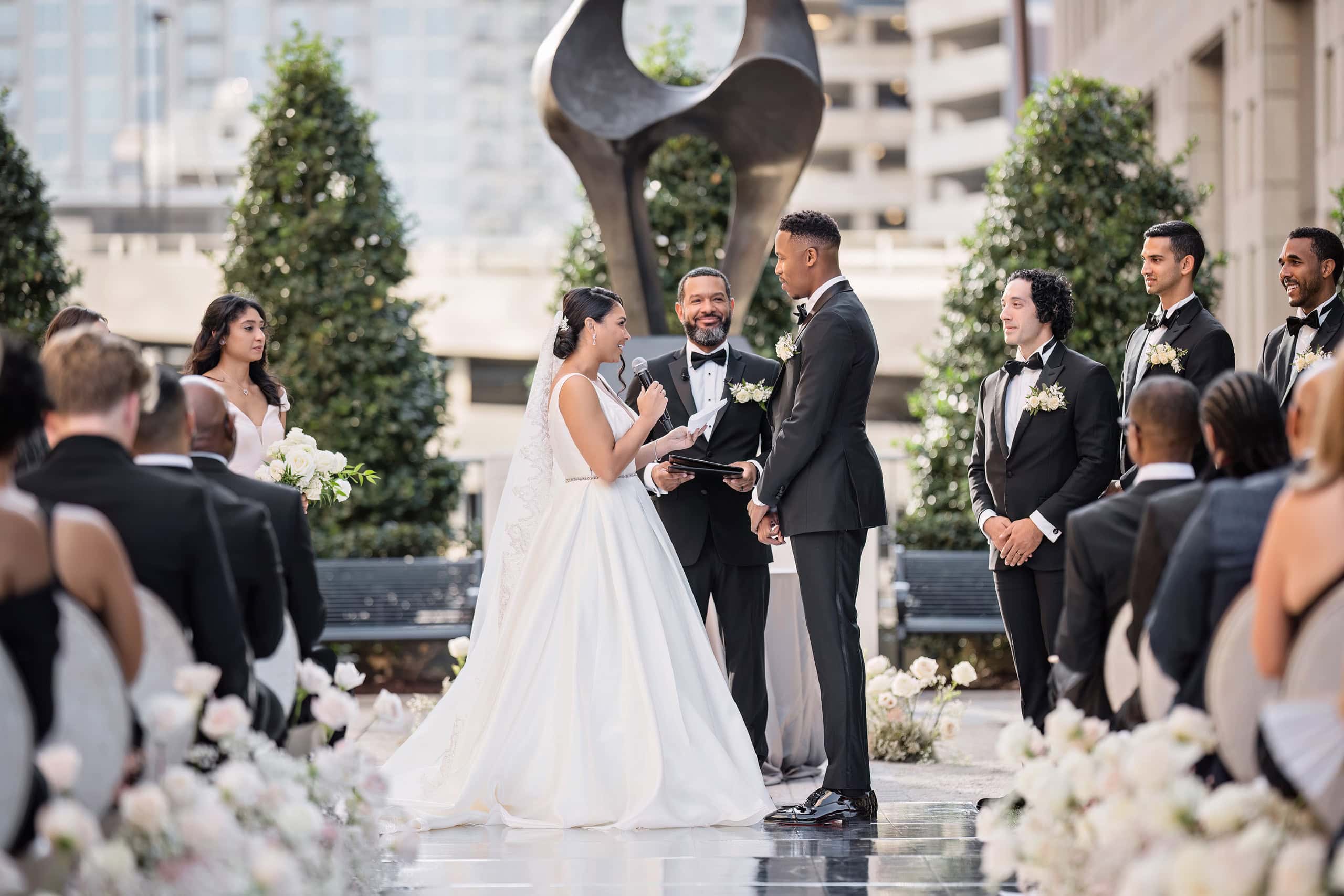 A bride and groom exchange vows at a Grand Bohemian Charlotte wedding ceremony in an urban setting, surrounded by groomsmen, bridesmaids, and guests with white floral arrangements and modern sculptures nearby.