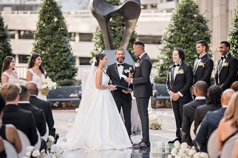 An outdoor wedding ceremony at the Grand Bohemian Hotel Charlotte featuring a bride and groom exchanging vows before a smiling officiant, surrounded by groomsmen, bridesmaids, and seated guests. A large