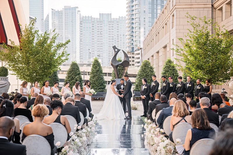 A wedding ceremony outdoors at the Grand Bohemian Hotel Charlotte, with a bride and groom at the altar, surrounded by guests seated in rows. The setting includes skyscrapers, trees, and a