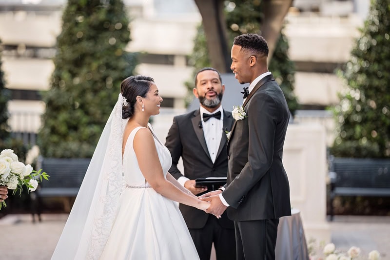 A bride and groom hold hands during their wedding ceremony at the Grand Bohemian Hotel Charlotte, with the officiant standing behind them in an outdoor setting adorned with white flowers.