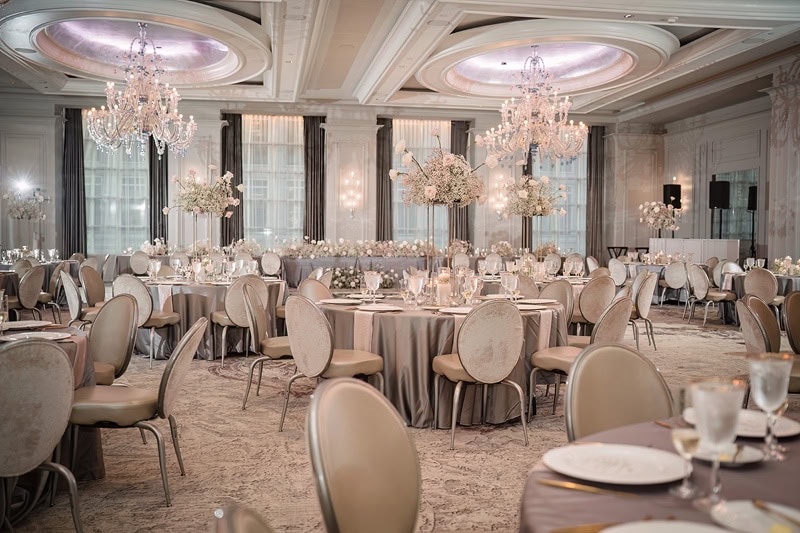 Elegant Grand Bohemian Hotel Charlotte banquet hall with grand chandeliers, round tables set with white linen, glassware, and floral centerpieces. Neutral tones and soft lighting create a luxurious atmosphere