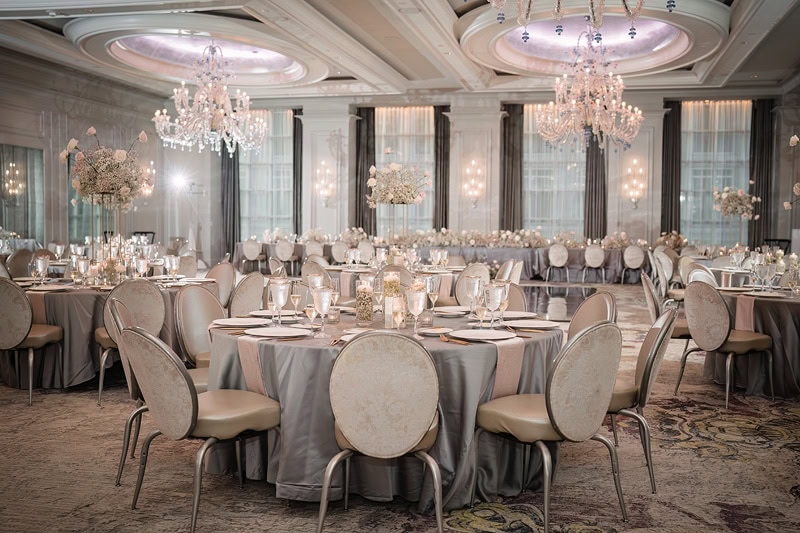Opulent banquet hall at the Grand Bohemian Hotel Charlotte set for a formal event, featuring round tables draped in grey cloths with gold-trimmed chairs. Crystal chandeliers and floral center
