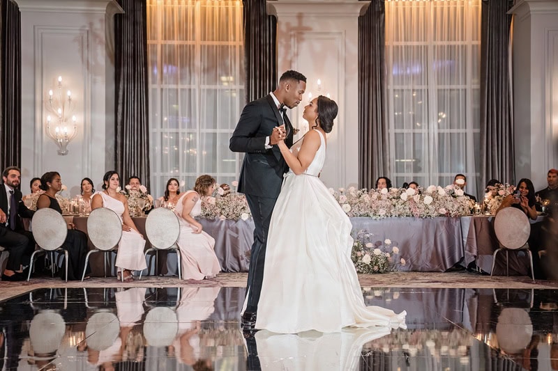 A bride and groom share a dance, surrounded by guests at an elegant wedding reception at the Grand Bohemian Hotel Charlotte. The ballroom features lavish decor with floral arrangements, chandeliers, and