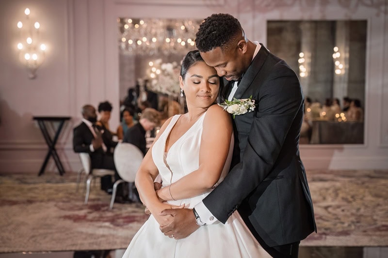 A newlywed couple shares an intimate dance in the elegant Grand Bohemian Hotel Charlotte, adorned with crystal chandeliers and twinkling lights, surrounded by wedding guests. The bride, in a white