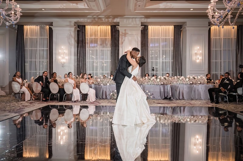 A newlywed couple shares a romantic dance on a glossy floor that reflects their image, surrounded by guests seated at elegant, floral-decorated tables in the luxurious Grand Bohemian Hotel Charlotte ballroom