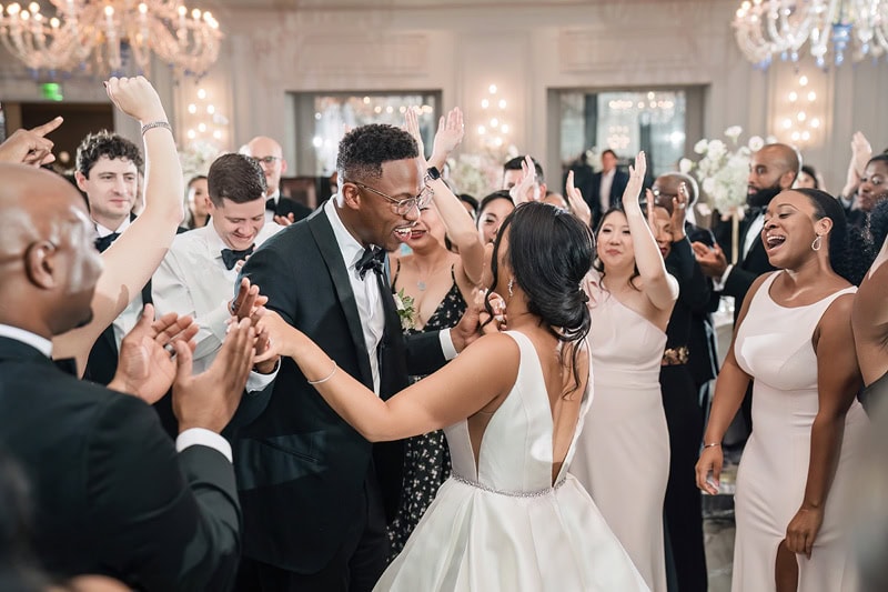A joyful bride and groom, dressed elegantly in a white gown and black tuxedo, dance through a cheering crowd of wedding guests at the Grand Bohemian Hotel Charlotte, in a well-l