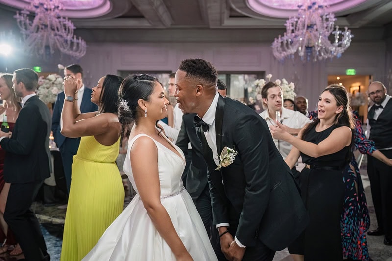 A joyful wedding reception scene at the Grand Bohemian Hotel Charlotte with a bride and groom kissing amidst dancing guests. Elegant chandeliers hang above. The bride in white and the groom in a suit