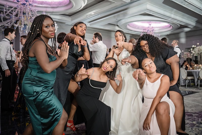A group of seven diverse women at a formal event, playfully posing and dancing at the Grand Bohemian Hotel Charlotte. They are variously dressed in elegant gowns and one in a white wedding