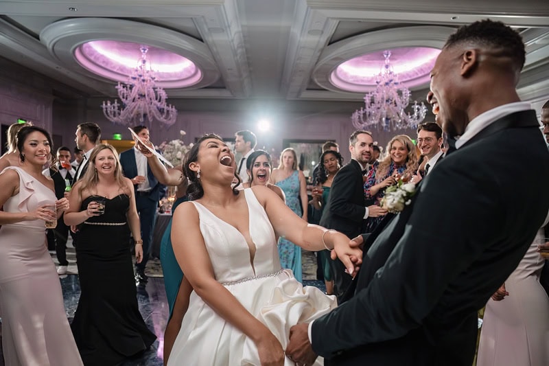 A joyful bride, wearing a white dress, laughs and reaches towards a groom in a black suit as they dance amidst guests at the Grand Bohemian Hotel Charlotte. The diverse crowd of guests is cl