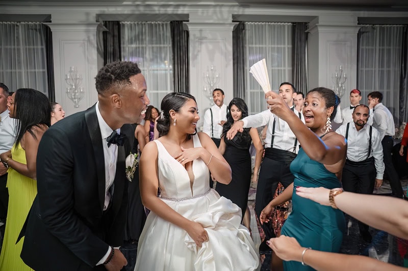 A joyful wedding reception scene at the Grand Bohemian Hotel Charlotte with a bride in a white dress and a groom in a black suit smiling at a woman in a teal dress who is enthusiastically showing them
