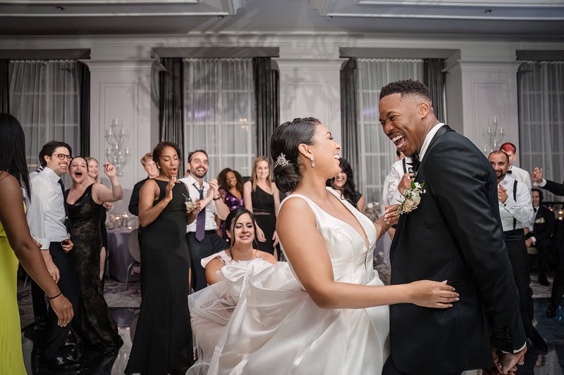 A joyful bride and groom dance at their wedding reception at the Grand Bohemian Hotel Charlotte. They are laughing together in a well-lit ballroom, surrounded by guests who are clapping and celebrating