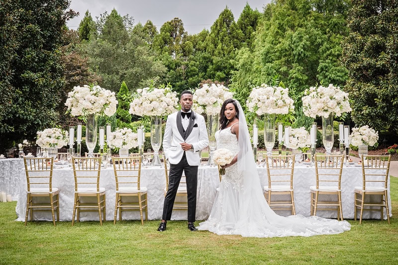 A bride and groom stand hand-in-hand outdoors at their wedding venue, a lush garden. They are elegantly dressed, he in a black one-button suit, she in a long white lace gown.