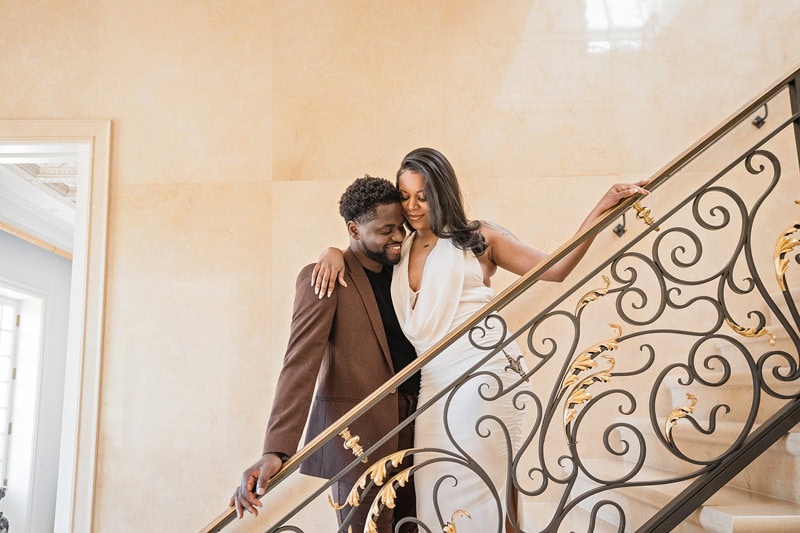 A couple descends a grand staircase at Oxbow Estate, the woman in a white dress leaning on the man in a brown jacket. They smile at each other, surrounded by an elegant interior with