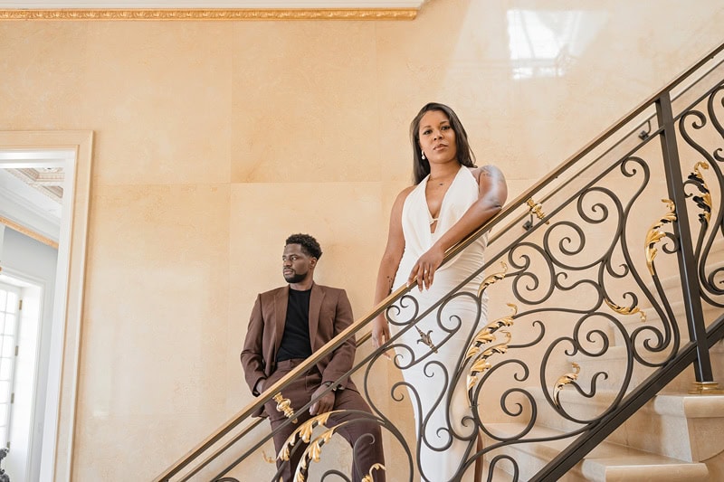 A woman in a white dress descends an ornate staircase dring their Oxbow Estate engagement session, holding the railing, gazing outward. A man in a brown suit sits on a step lower down,