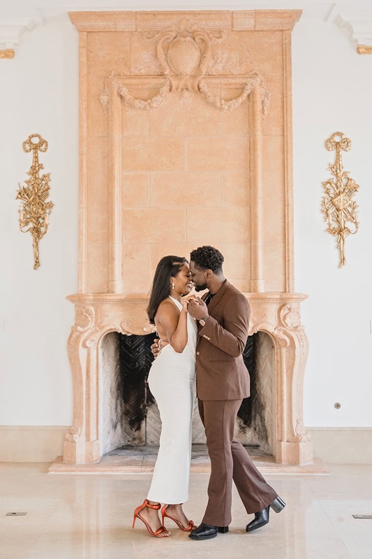 An elegant couple dances intimately during their Oxbow Estate engagement photos by an ornate fireplace with a detailed golden plaque and sconces. The woman is in a long white dress and red heels, the man