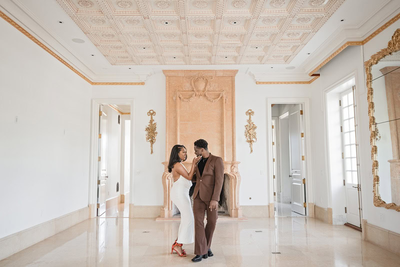 A couple stands close together in the luxurious Oxbow Estate hall with ornate walls and a detailed ceiling. The man in a brown suit kisses the woman's forehead as she, dressed in white pants