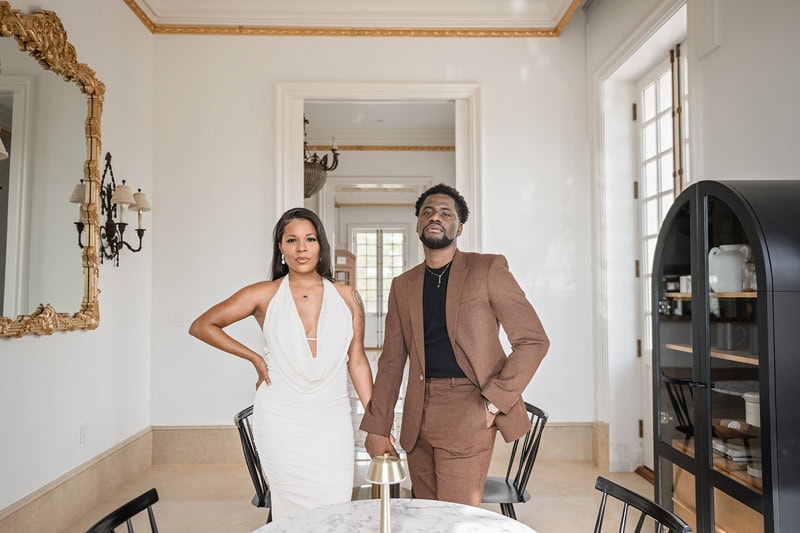 A man and a woman stand confidently in an elegant room with classic decor at Oxbow Estate. The man wears a brown suit while the woman is in a white dress. A round black table and