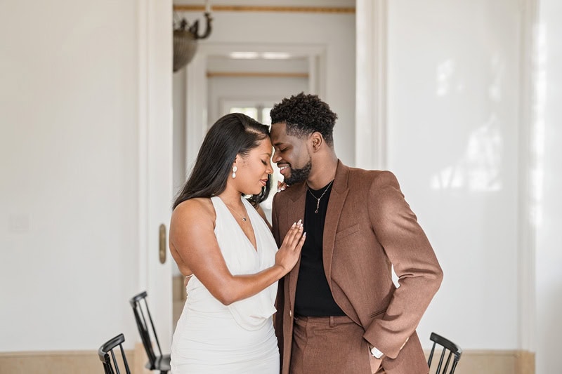 A couple affectionately touching foreheads and holding hands in a warmly lit corridor during their Oxbow Estate engagement photos, the woman in a white dress and the man in a brown suit jacket.