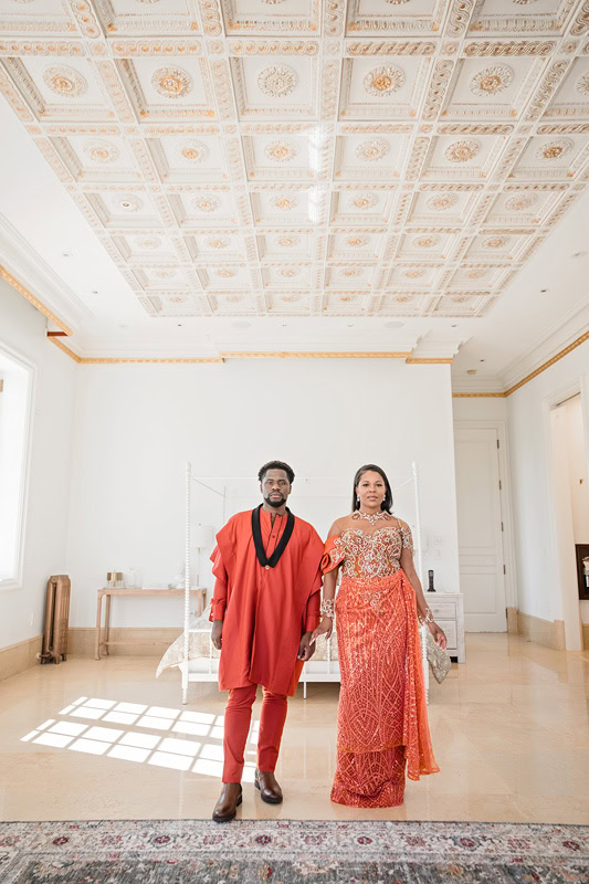 A couple dressed in traditional attire stand hand in hand at Oxbow Estate, with intricate ceiling patterns, a polished floor, and soft natural light enhancing their engagement photos. The man wears a vibrant red