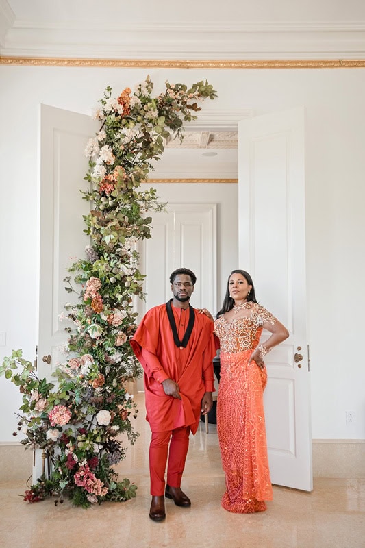 A man and woman pose elegantly in a luxurious room at Oxbow Estate. The man, in a vibrant red traditional outfit, stands beside the woman dressed in an ornate orange gown. A