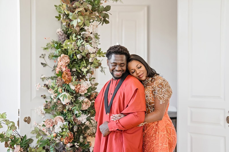A joyful couple embraces at Oxbow Estate, with the woman leaning her head on the man’s shoulder. Both wear traditional, ornate outfits, the man in red and the woman in orange,