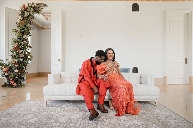 A joyful couple sits closely on a white sofa at Oxbow Estate, with the man in a red outfit whispering into the smiling woman's ear. She is wearing a richly detailed orange sare