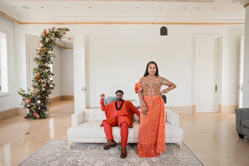 A black couple in striking traditional attire poses at Oxbow Estate. The woman, in a detailed orange dress, stands confidently, while the man in a vibrant red suit sits on a white couch,