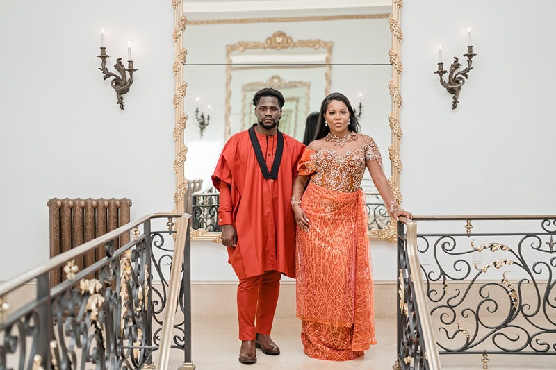 A couple in traditional Nigerian attire stand confidently on an ornate stairway at Oxbow Estate. The man wears a vibrant red agbada with embroidery, while the woman is in an intricately