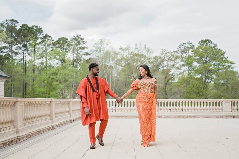 A man and a woman happily walk hand in hand on a stone bridge at Oxbow Estate. Both wear traditional orange attire, with the man in an embroidered robe and the woman in a detailed gown